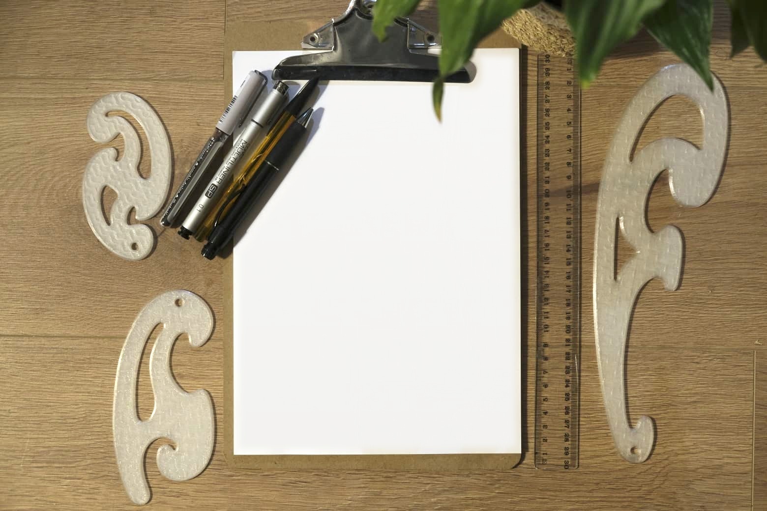 A blank sheet of paper on a clipboard, with fineliners, a pencil and pen, a ruler and French curves laid out around it